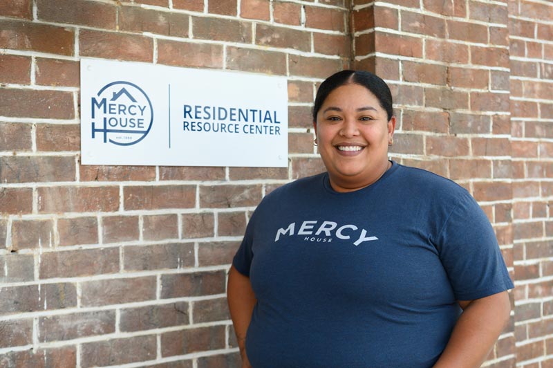 Ermila Ermila Headshot, woman standing in front of a brick wall and a sign reading "Mercy House residential resource center."