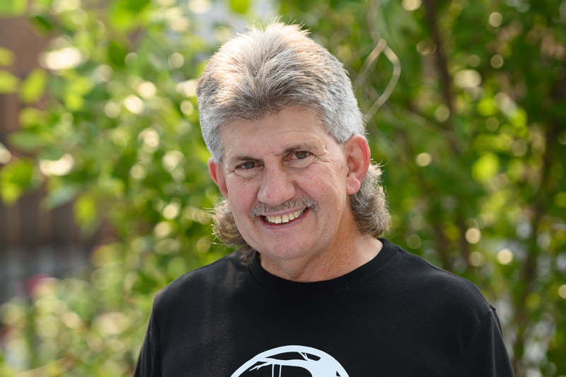 Steve Steve headshot, man standing in front of a green leafy tree and black fence